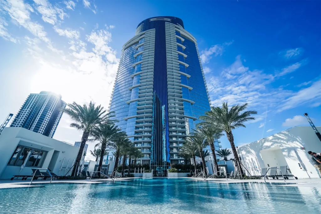Blue glass skyscraper at Paramount Miami Worldcenter rises behind a swimming pool, palm trees, and a terrace with one person visible