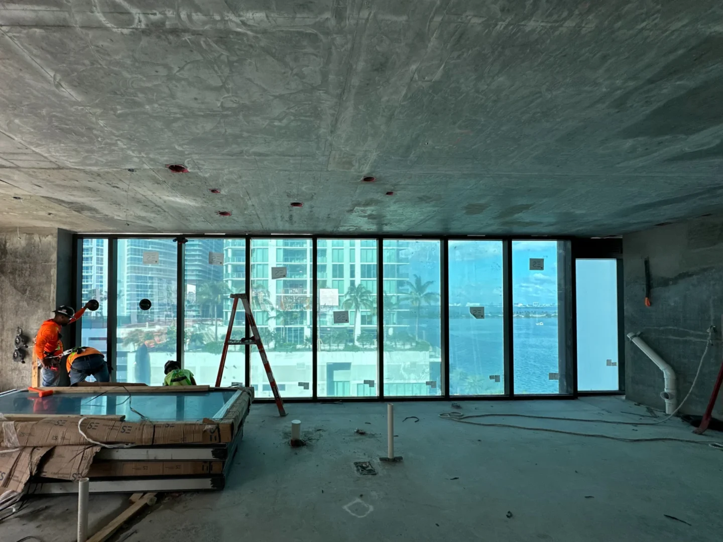 A concrete ceiling covers an unfinished room featuring large windows overlooking water and construction workers near a ladder