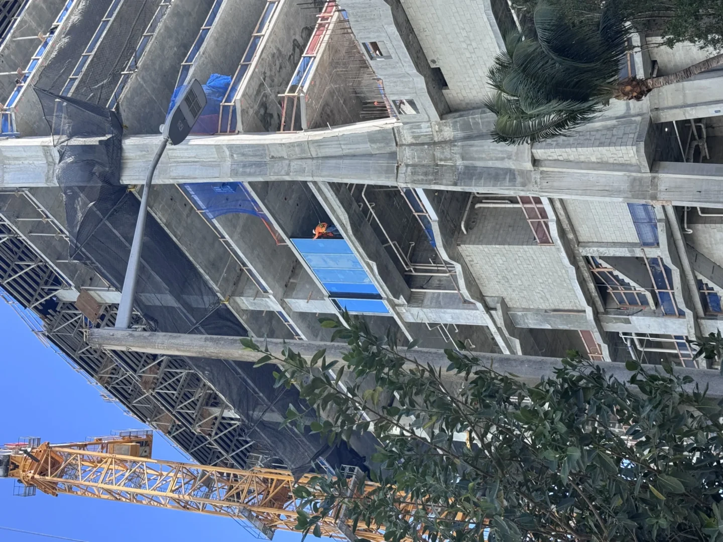 An unfinished concrete building features blue scaffolding, black safety netting, and a yellow crane near green trees