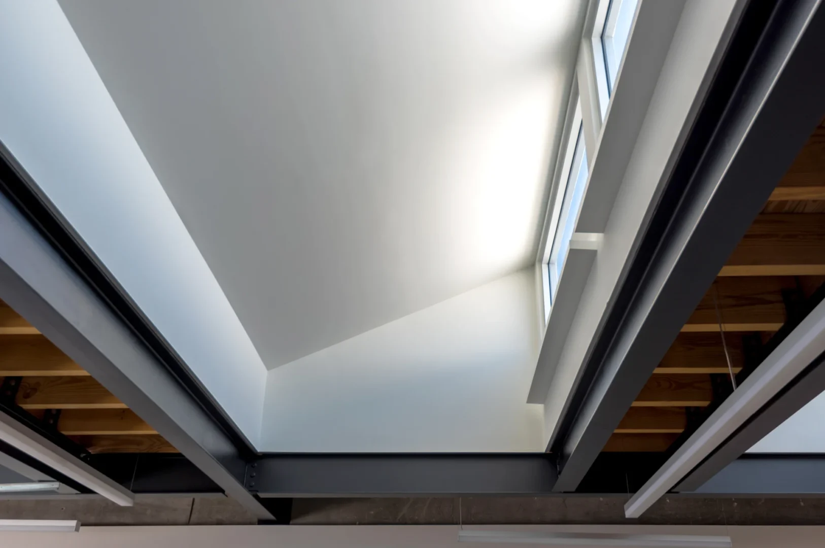 Dark steel beams and wooden planks frame a brightly lit ceiling and window in the ODP Architecture & Design Headquarters
