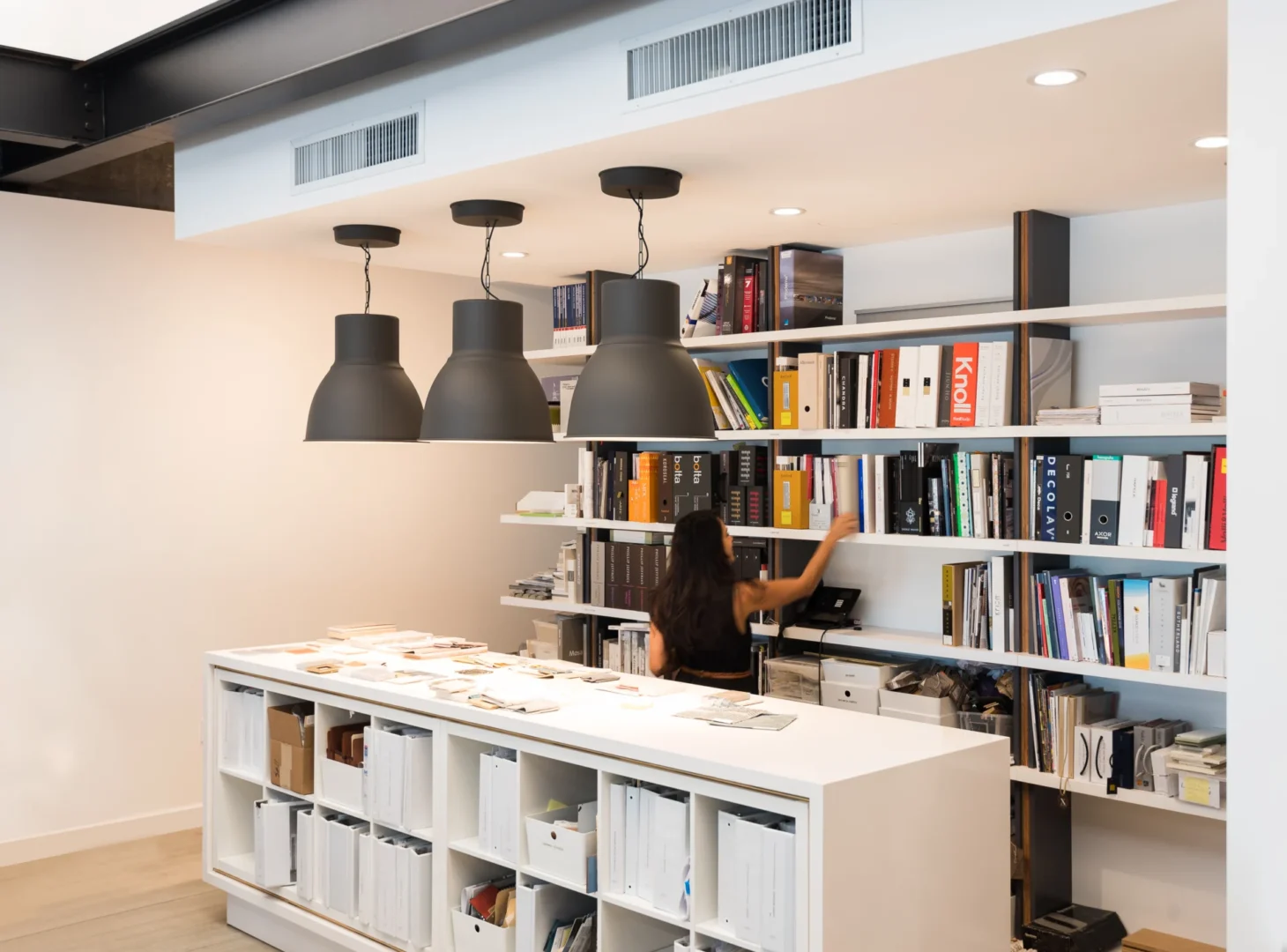 Three black pendant lights hang above a person reaching for books on shelves in the ODP Architecture & Design Headquarters workspace