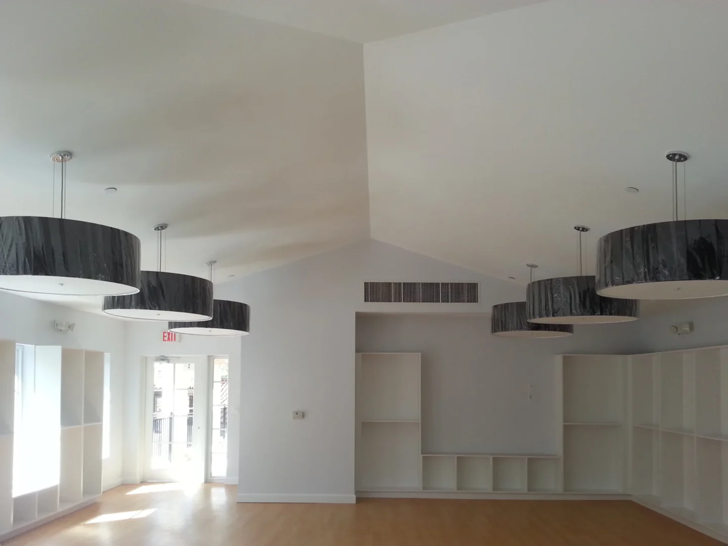 Large, dark drum pendant lights hang from a high ceiling above white built-in shelving in a room at Fisher Island Day School