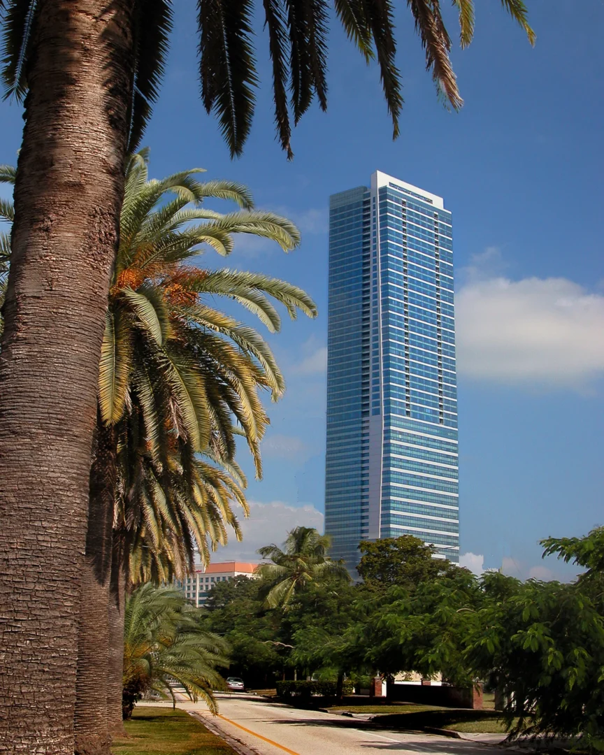 Large palm tree trunks stand beside a blue glass skyscraper at Naples Beach Four Seasons Resort & Residences