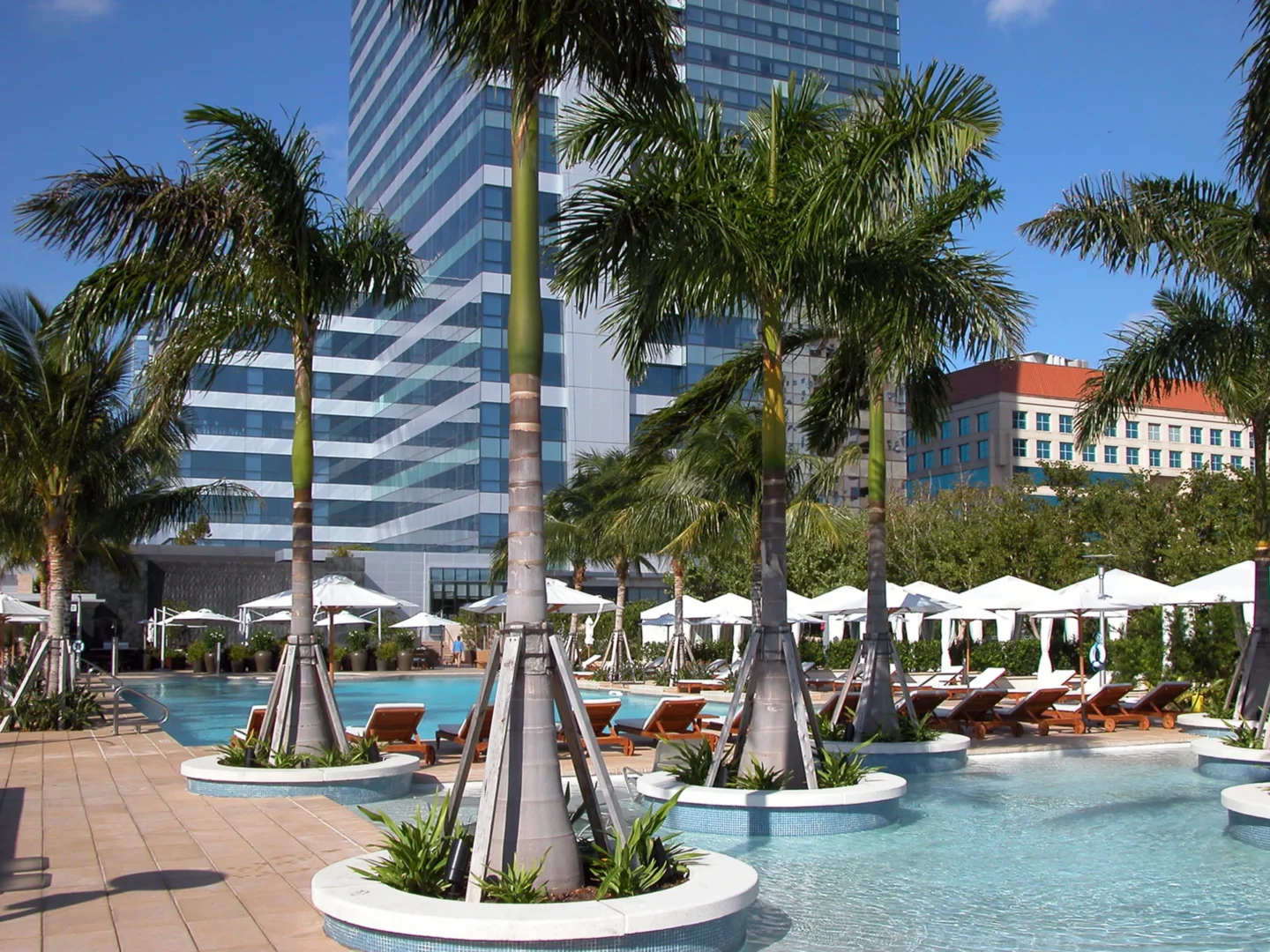 A tall glass skyscraper stands behind palm trees and a pool terrace at the Naples Beach Four Seasons Resort & Residences