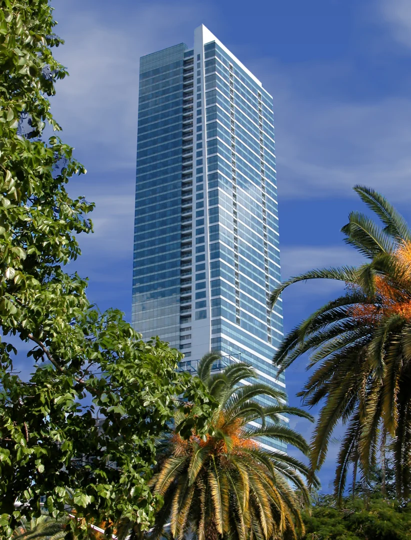 A blue glass skyscraper for Naples Beach Four Seasons Resort & Residences rises behind green trees and palm trees under a blue sky