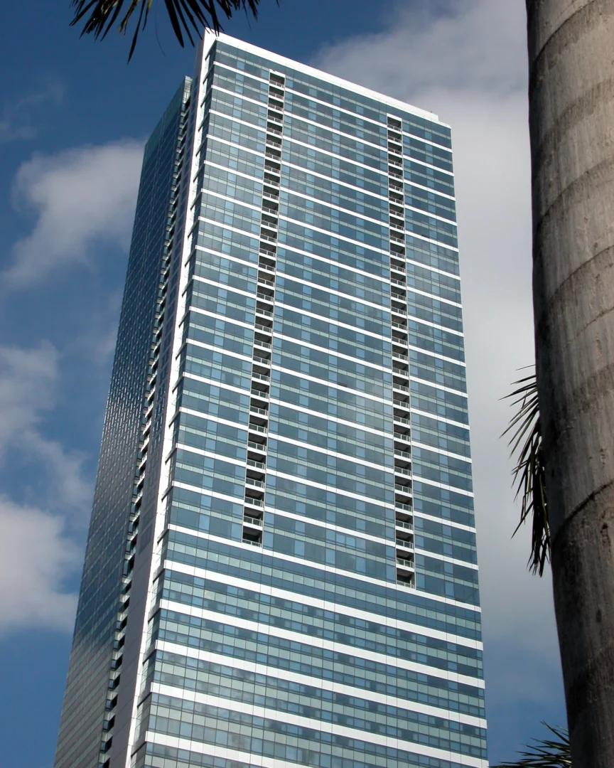 A blue glass high-rise for Naples Beach Four Seasons Resort & Residences stands against a cloudy sky with palm leaves visible