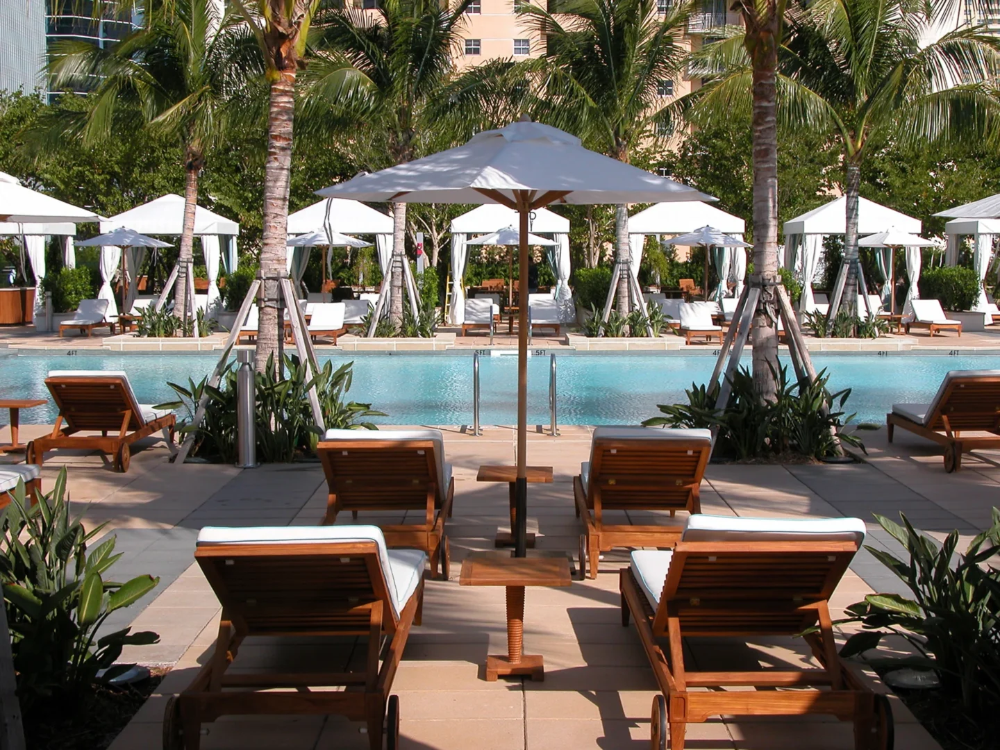 Wooden lounge chairs face a swimming pool with white tents and palm trees at Naples Beach Four Seasons Resort & Residences