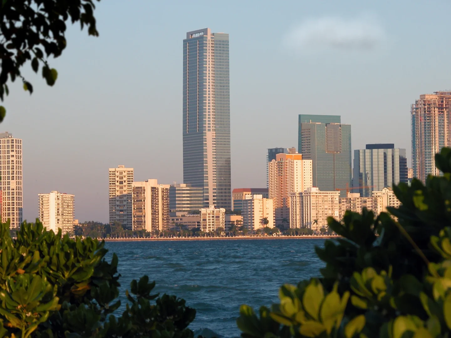 Green foliage frames skyscrapers across blue water for the Naples Beach Four Seasons Resort & Residences