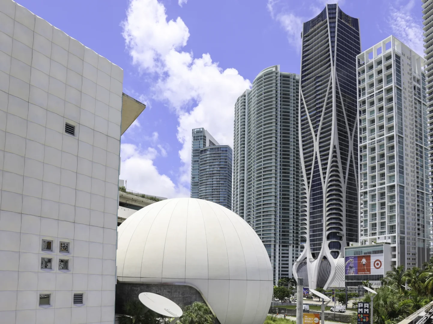Skyscrapers including One Thousand Museum rise beside a large white dome under a blue sky with clouds