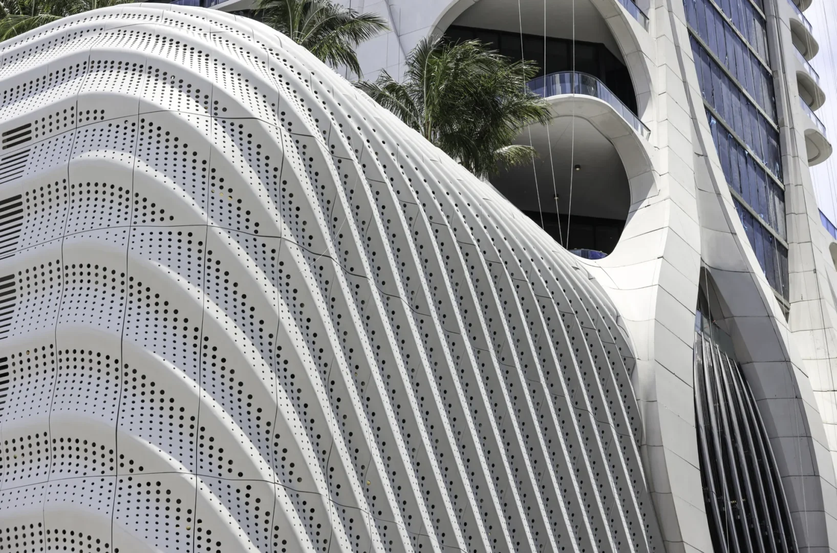Perforated white curved structure of the One Thousand Museum alongside glass windows, palm trees, and terraces