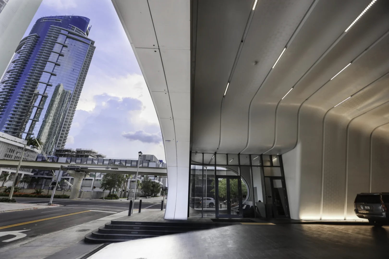 A curved white architectural overhang from One Thousand Museum covers a driveway and vehicle near a blue glass skyscraper