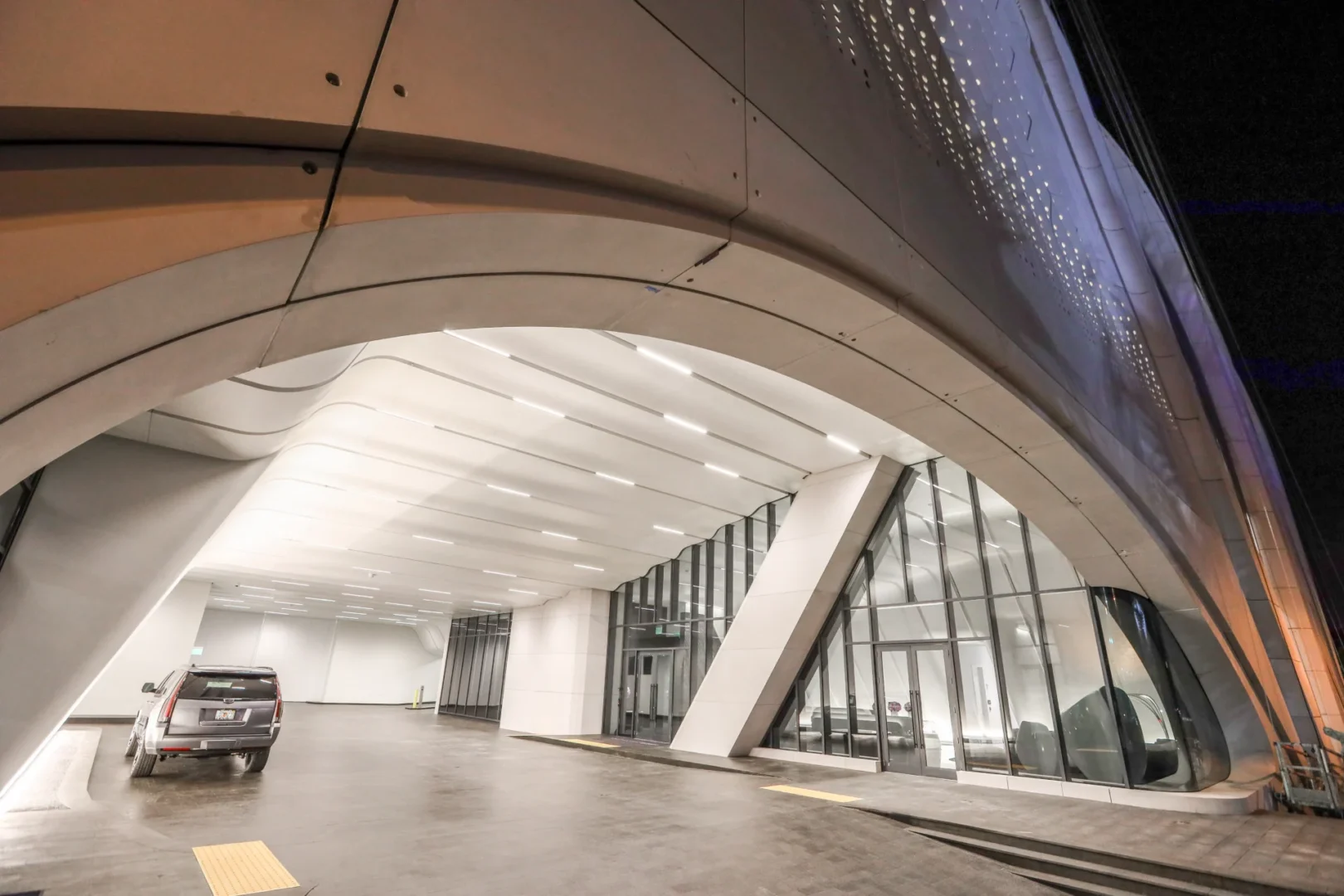 A large concrete arch at One Thousand Museum covers an illuminated driveway with a parked silver SUV and glass walls