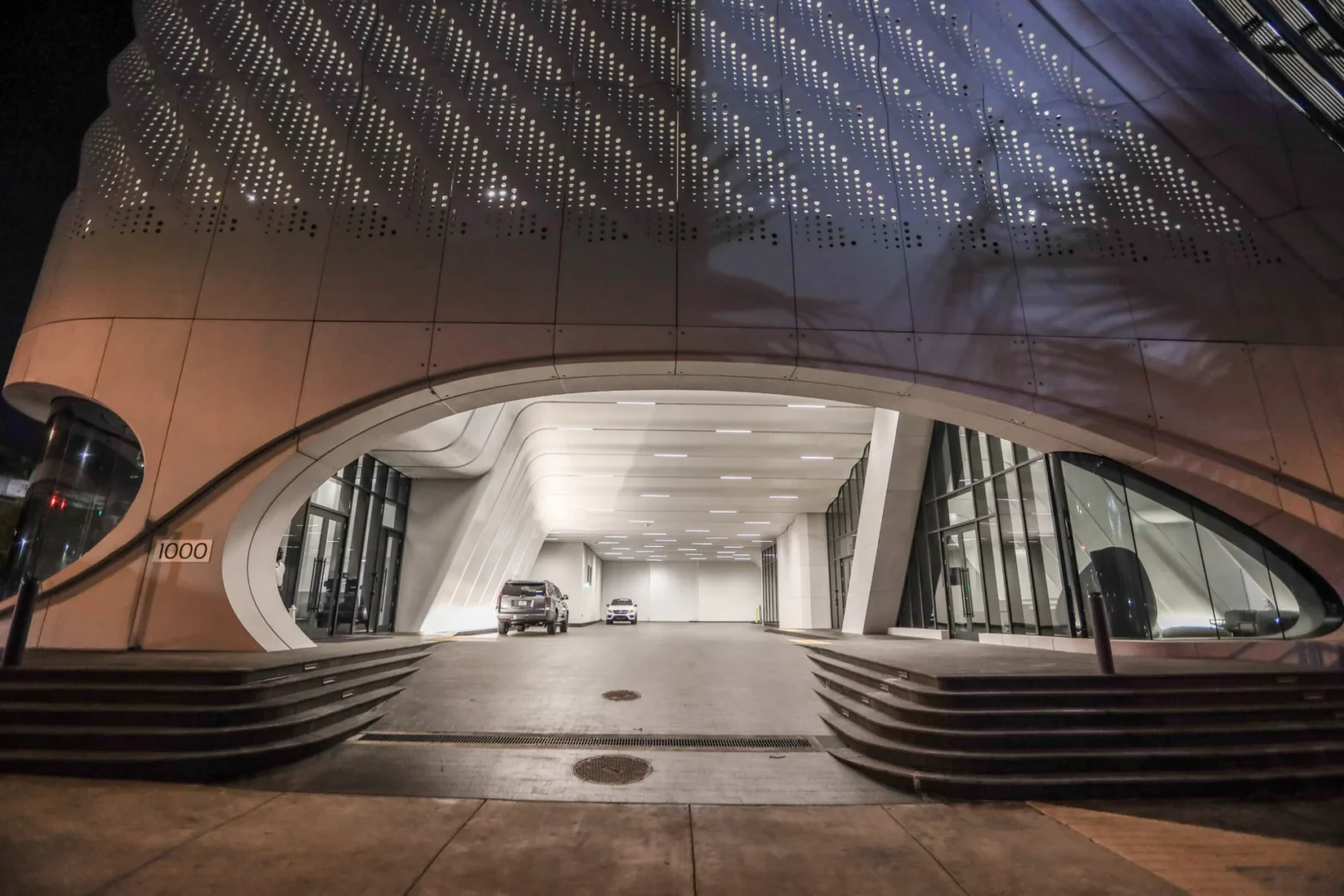 A white, perforated facade of One Thousand Museum arches over a driveway containing two cars