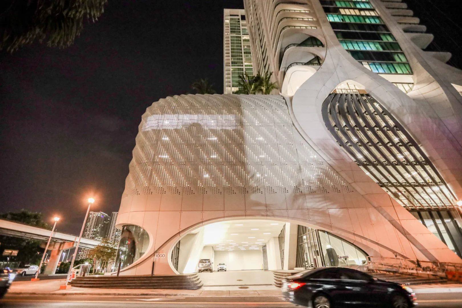 White, curved-concrete sections of One Thousand Museum feature a perforated facade above a street at night near a blurred car