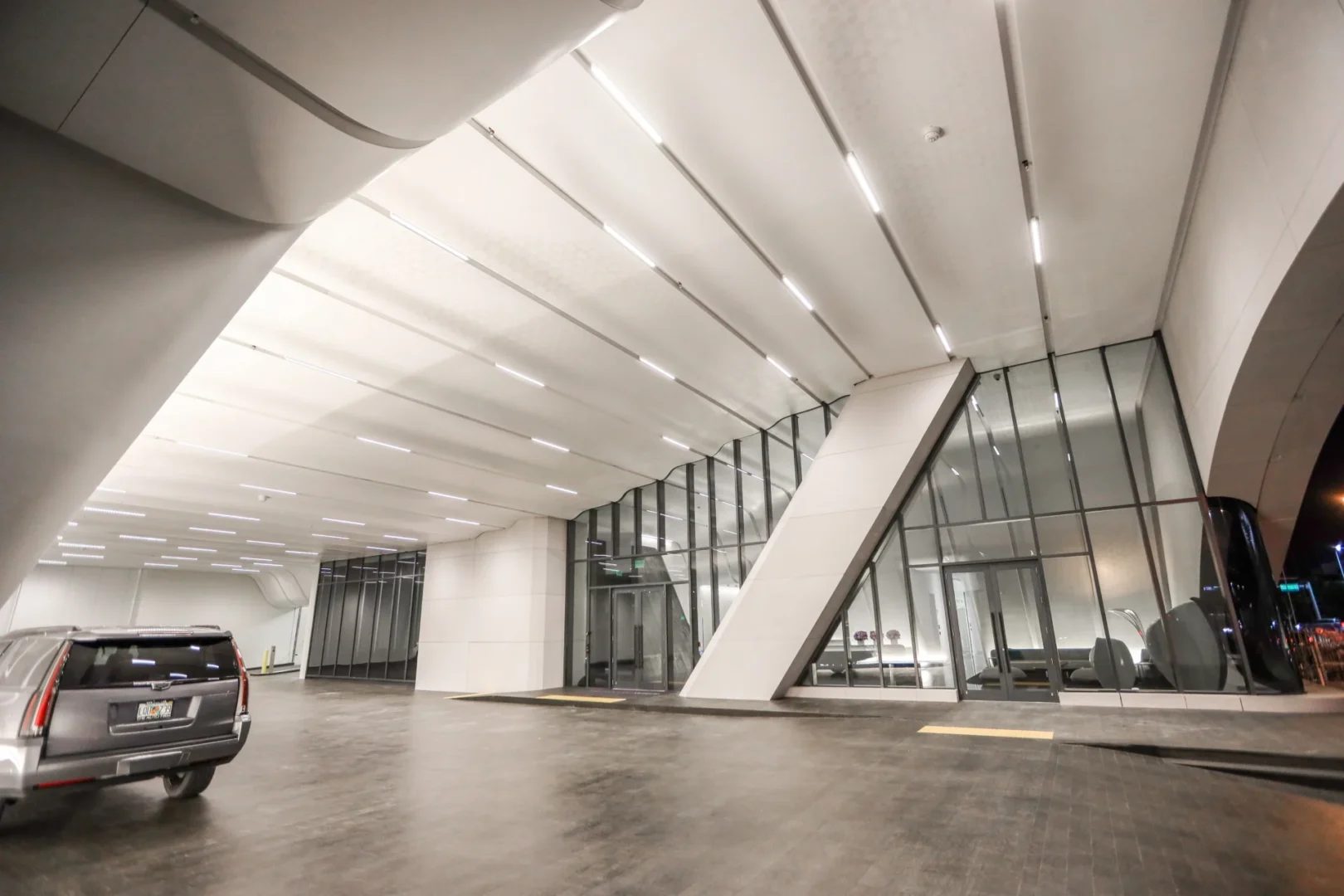 An illuminated white architectural ceiling at One Thousand Museum covers glass walls and a driveway with a silver SUV