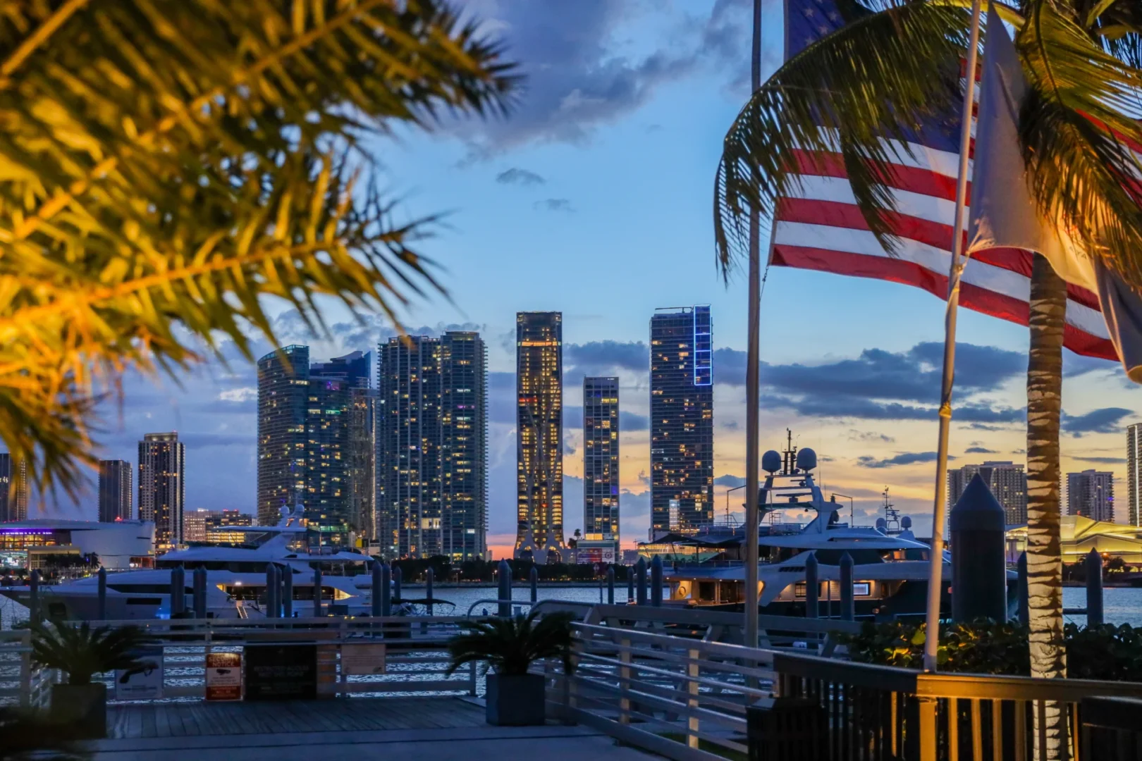 Palm tree leaves and an American flag border a skyline including One Thousand Museum and yachts docked in a marina at dusk