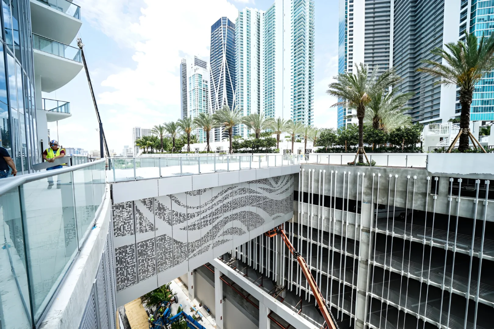 Un puente peatonal con motivos decorativos en Paramount D-East se extiende por encima de un aparcamiento, cerca de una persona con chaleco de seguridad que se encuentra en una terraza adyacente.
