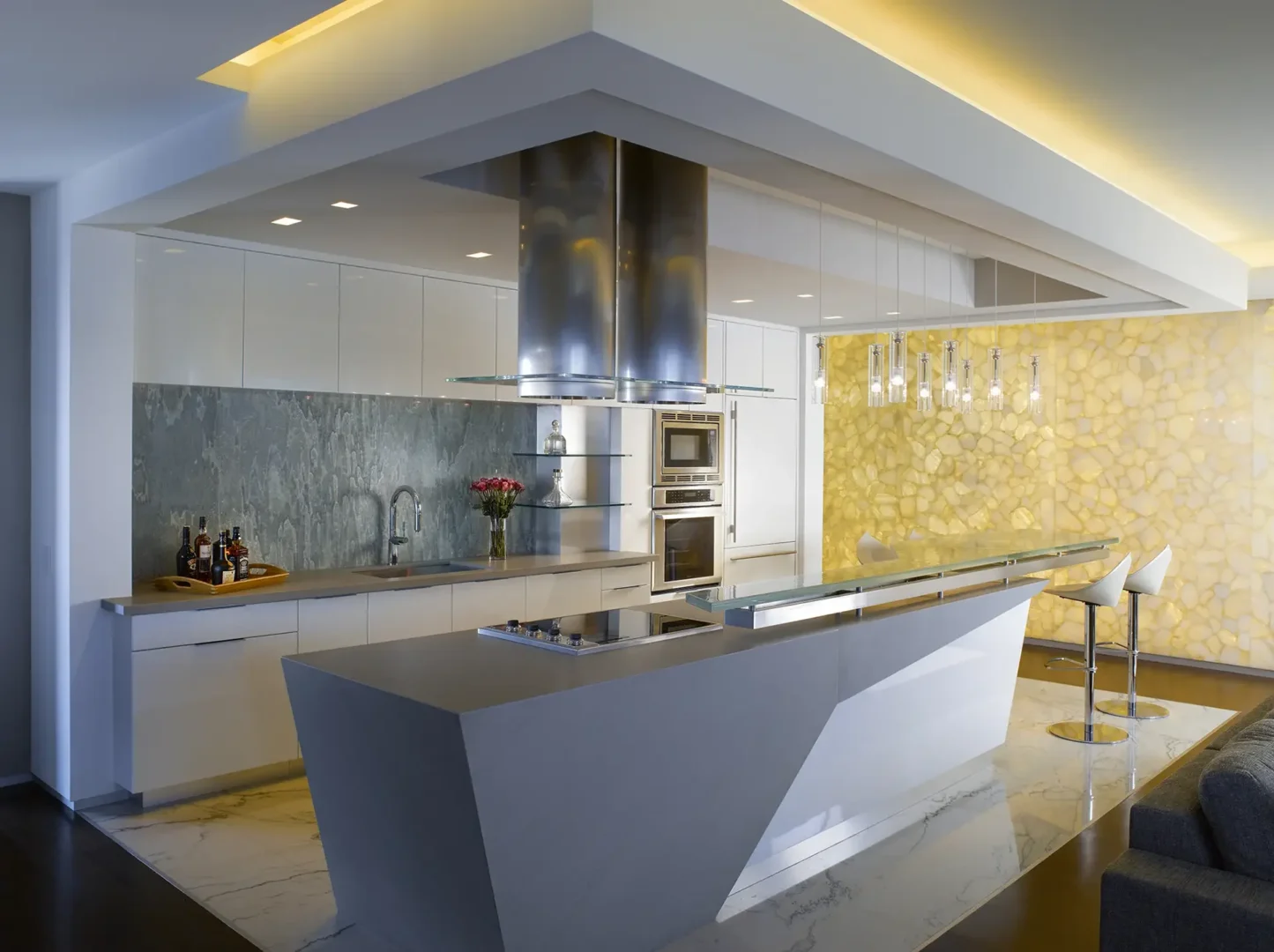A stainless steel vent hood hangs above a grey kitchen island and white cabinetry in Penthouse 1 near a backlit stone wall