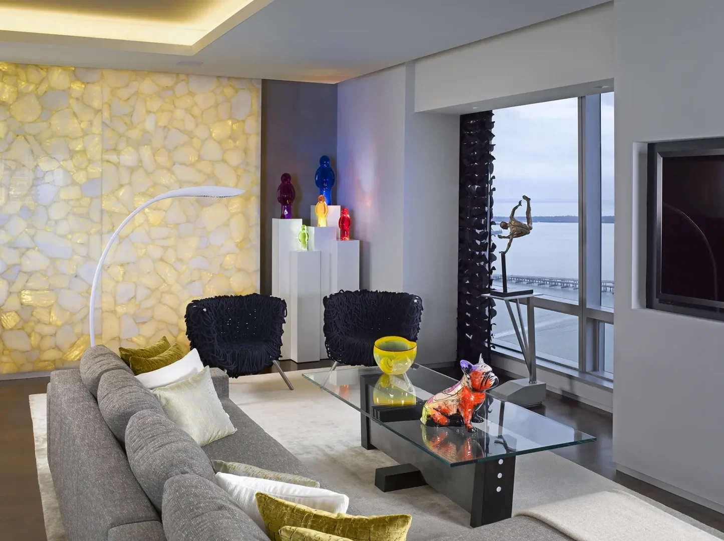An illuminated textured wall is in the Penthouse 1 living room with a grey sofa, glass coffee table, and window view of water