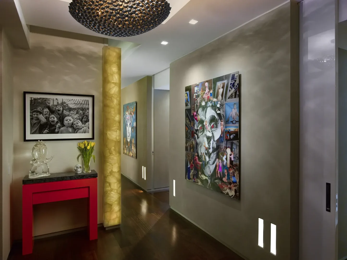 A large dark textured light fixture hangs over a hallway in Penthouse 1 containing artwork and a red console table