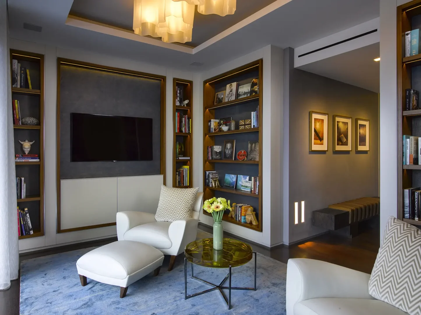 White armchairs and a glass coffee table sit on a blue rug beside built-in bookshelves and a television in Penthouse 1