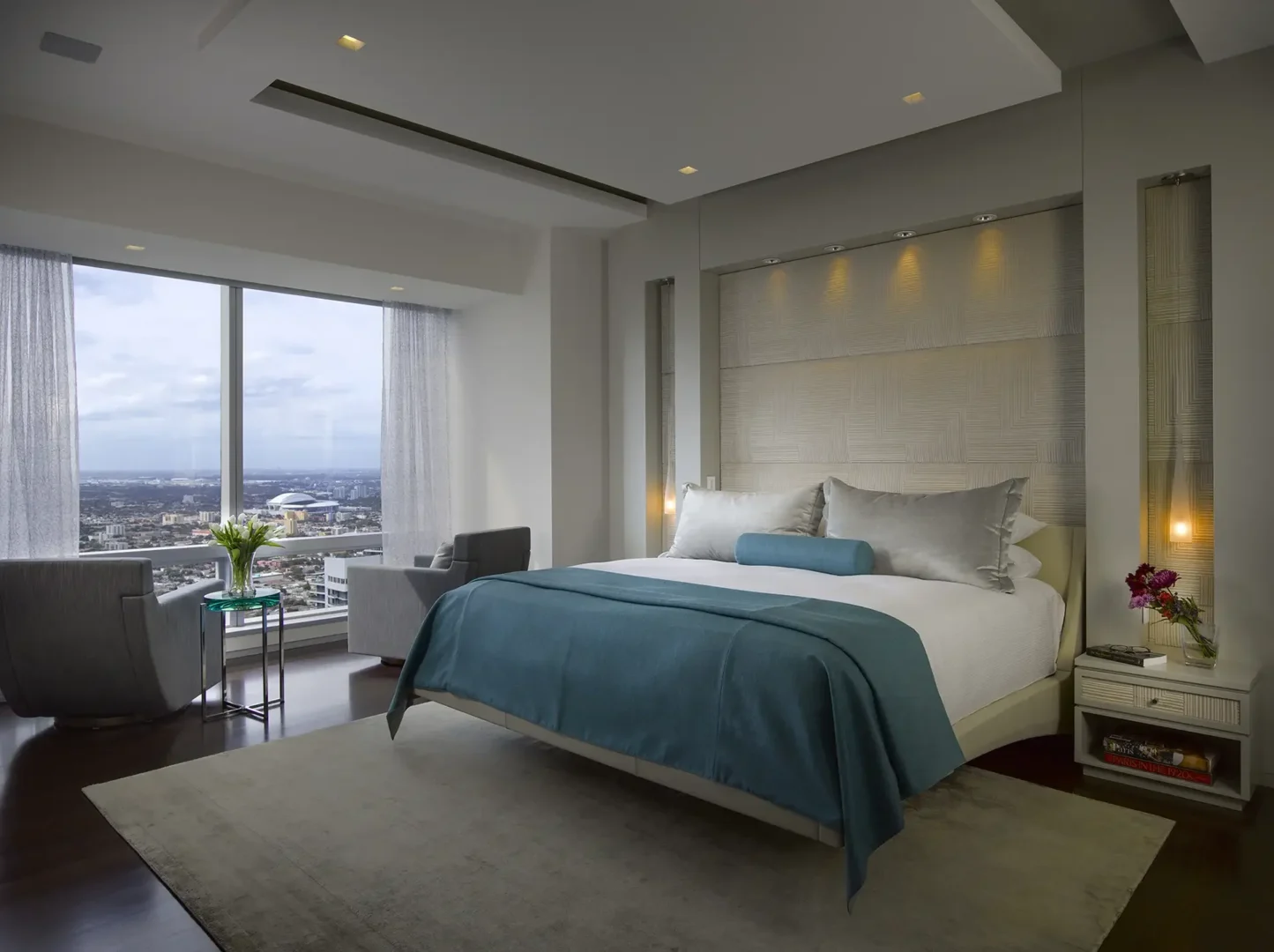 A bed with a blue throw sits in the Penthouse 1 bedroom alongside an armchair and a large window overlooking a city view