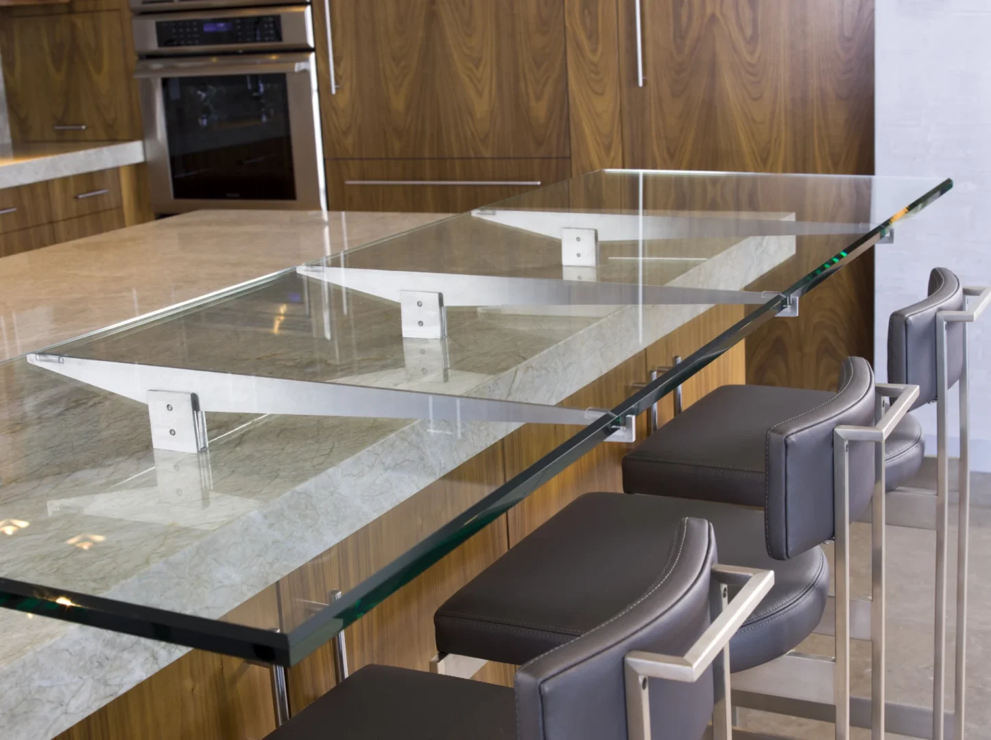 La prolongación con encimera de cristal de una barra de mármol del ático 3 cuenta con muebles de madera y taburetes de bar grises.