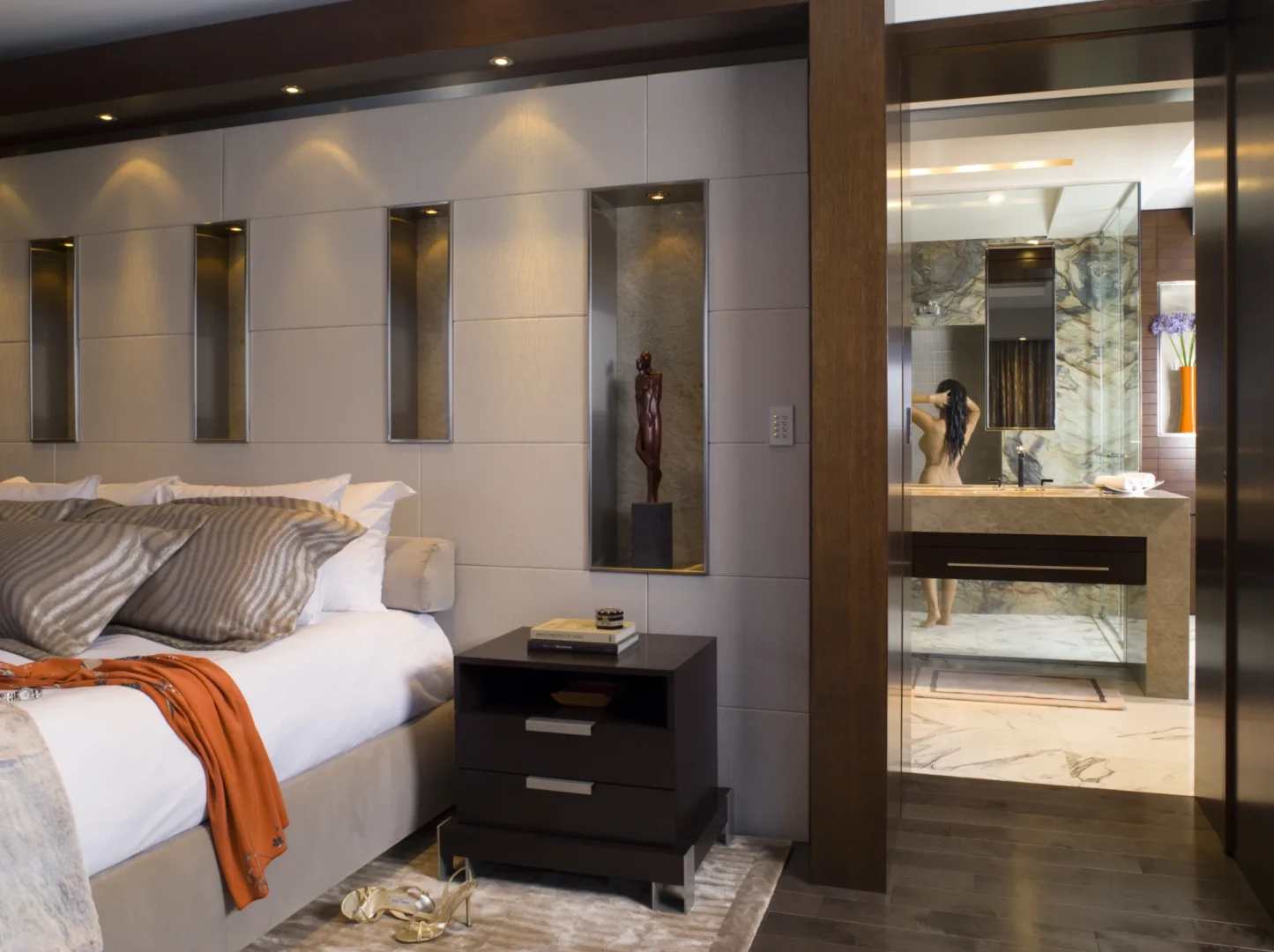 En un ático de tres dormitorios hay una cama y una mesita de noche oscura junto a la puerta abierta de un cuarto de baño, donde hay una persona de pie.