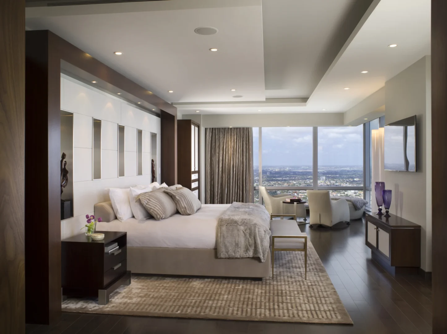 En un ático de tres dormitorios, con amplios ventanales que dan al perfil urbano, hay una cama con sábanas y almohadas blancas.