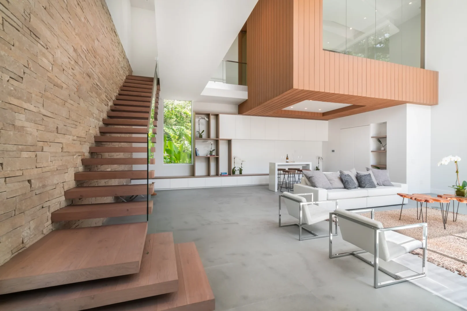 Wooden stairs along a stone wall are adjacent to an open kitchen and living area in the South Miami Residence