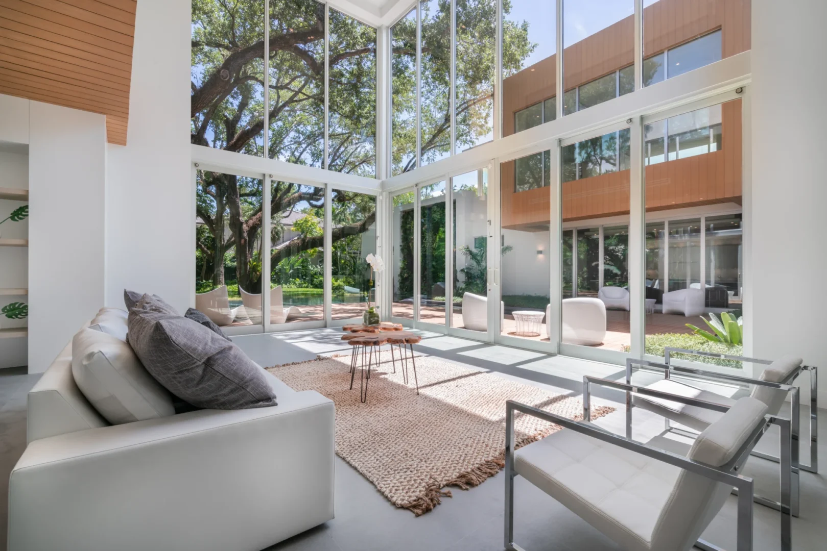 A white sofa and chairs in South Miami Residence face large glass windows overlooking trees and a wooden building structure