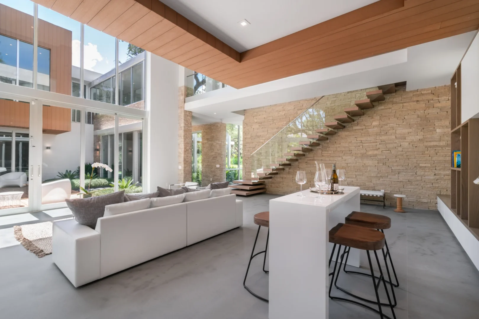 A white sectional sofa and kitchen island in the South Miami Residence are near a stone staircase and large glass walls