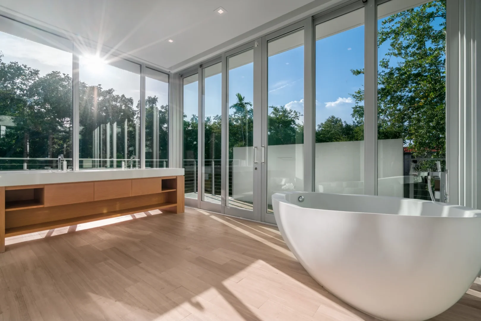 A white freestanding bathtub sits beside a wooden vanity in the South Miami Residence bathroom with large glass windows