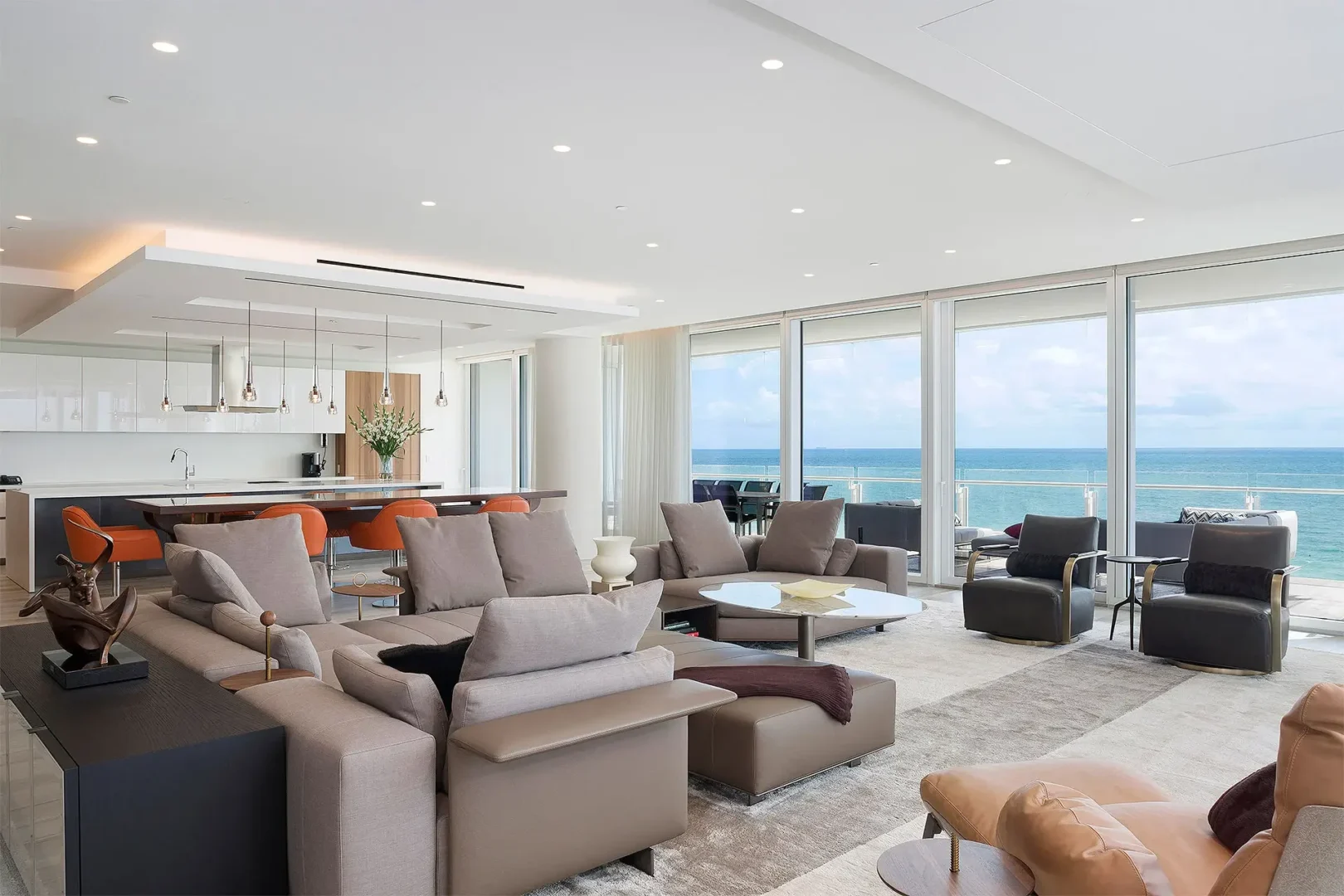 Grey sofas and armchairs in the Surfside Seaway lounge area face windows overlooking the ocean near a kitchen and dining space