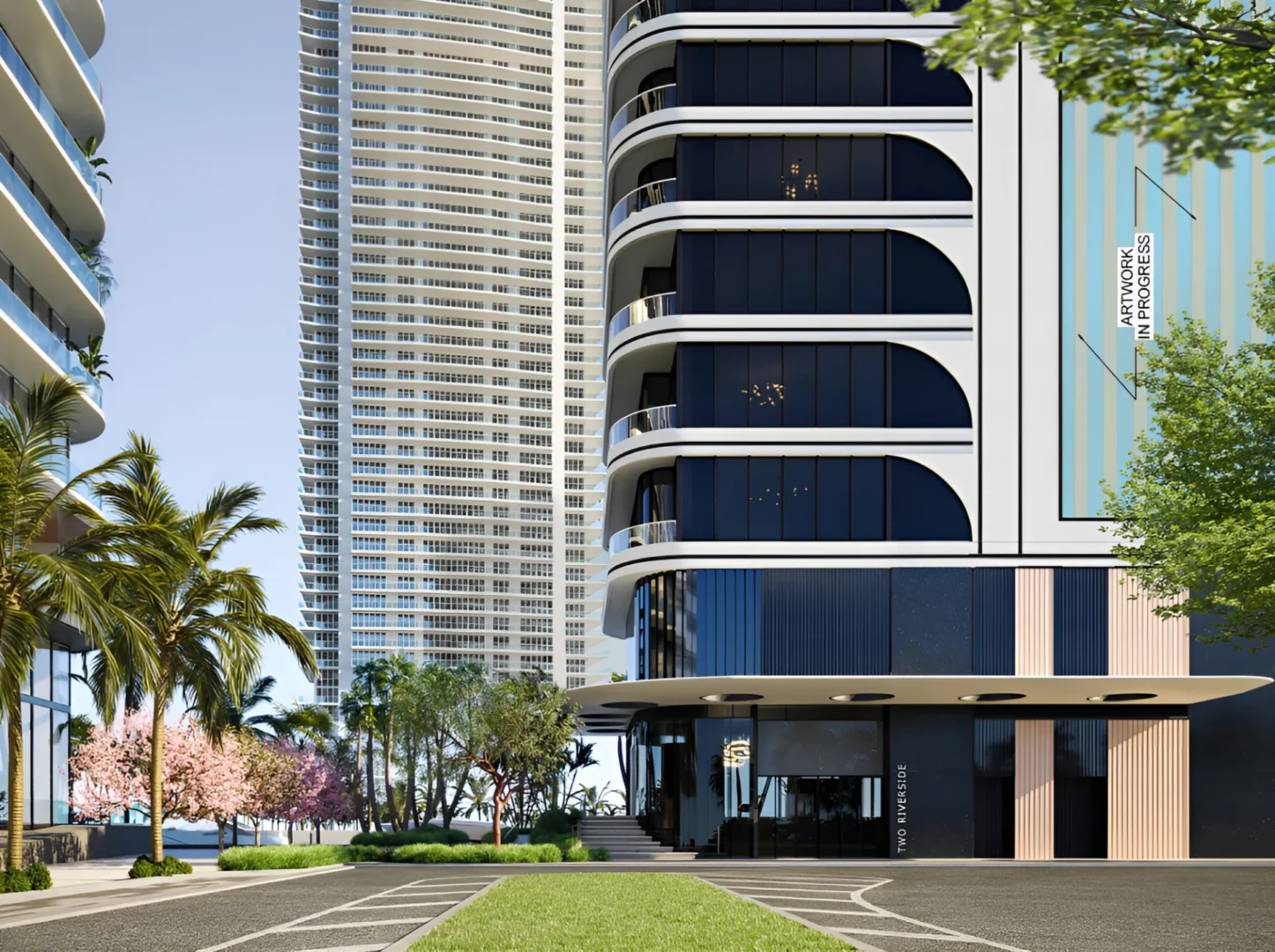 A white and dark blue building of Two Riverside stands next to a tall skyscraper, palm trees, and flowering trees near a roadway