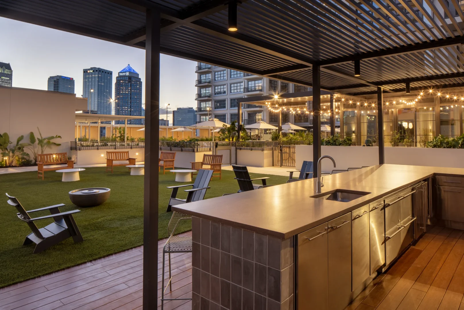 Una cocina al aire libre con encimera metálica y fregadero en la terraza del Asher Water Street de Tampa, con vistas al césped y al perfil urbano de la ciudad.