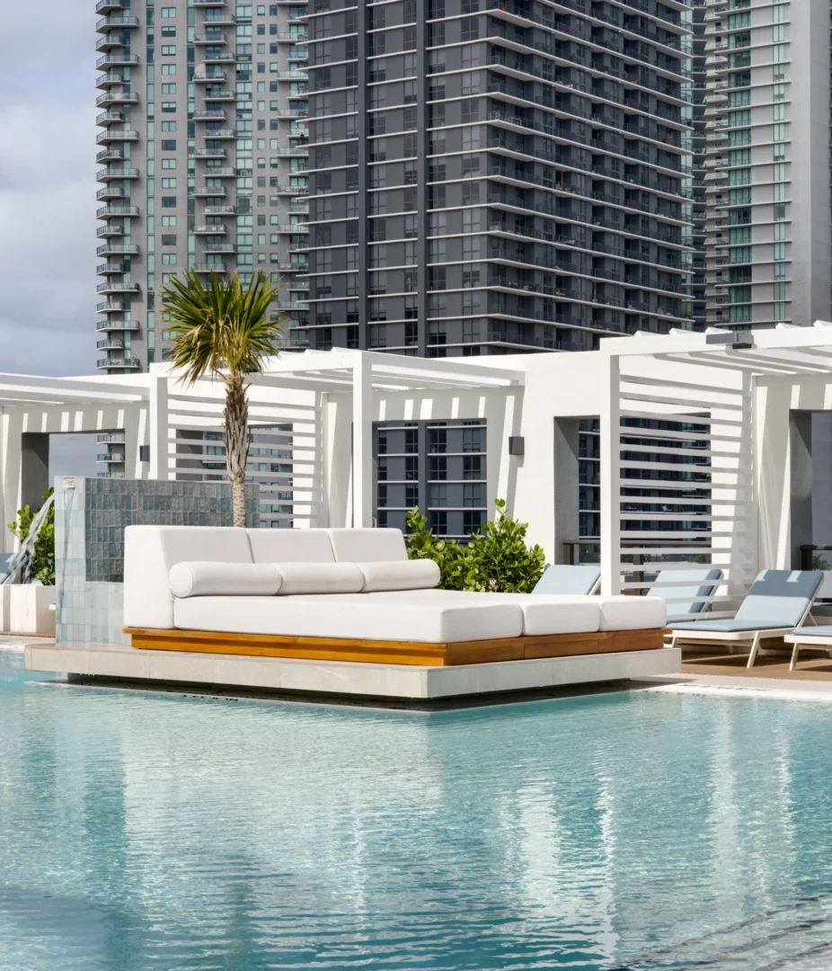 A large white daybed on a platform sits in a swimming pool within the Forma project, backed by high-rise buildings
