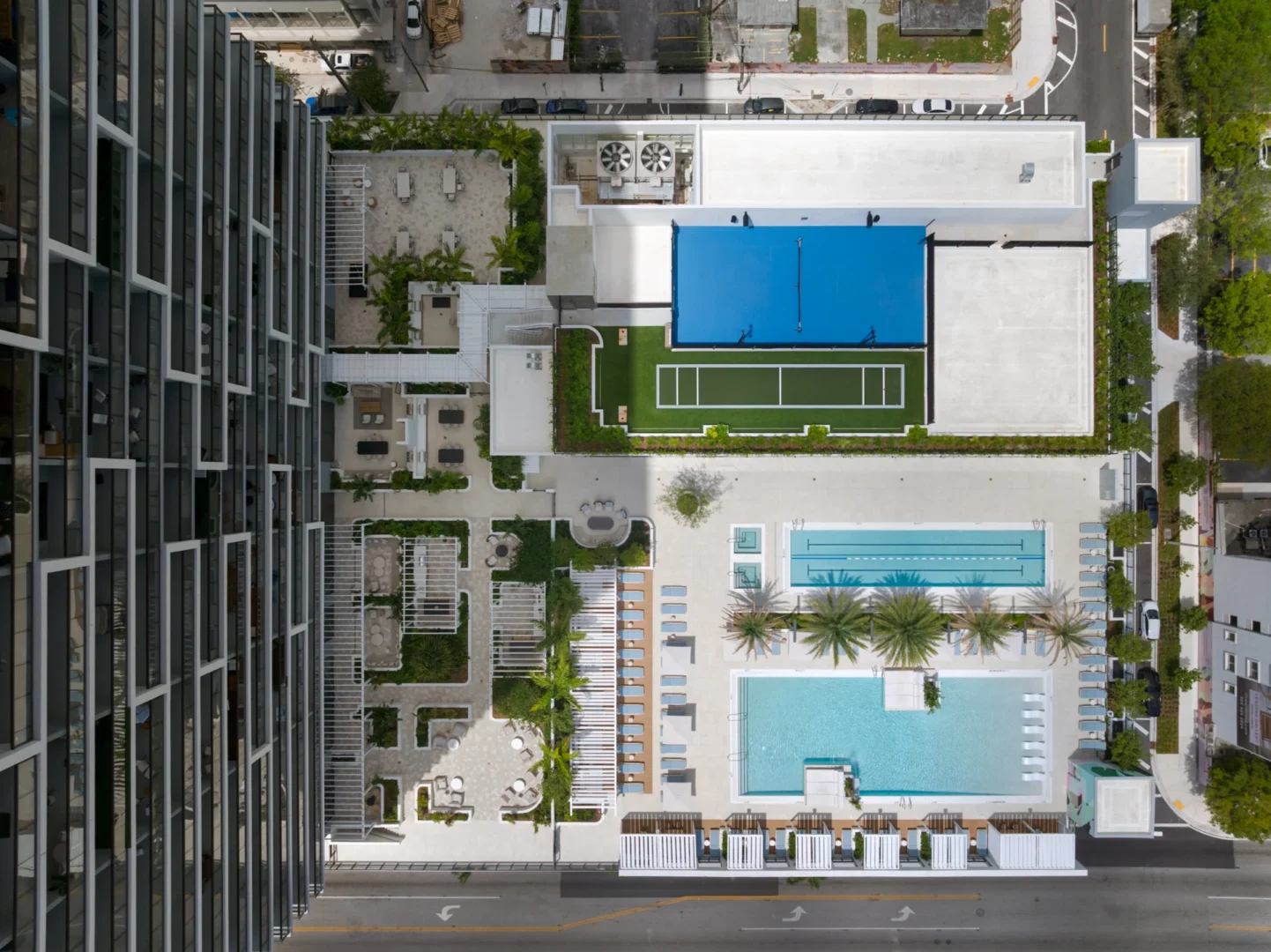 A high-rise building casts shadows over the tennis courts, swimming pools, and outdoor seating areas at Forma