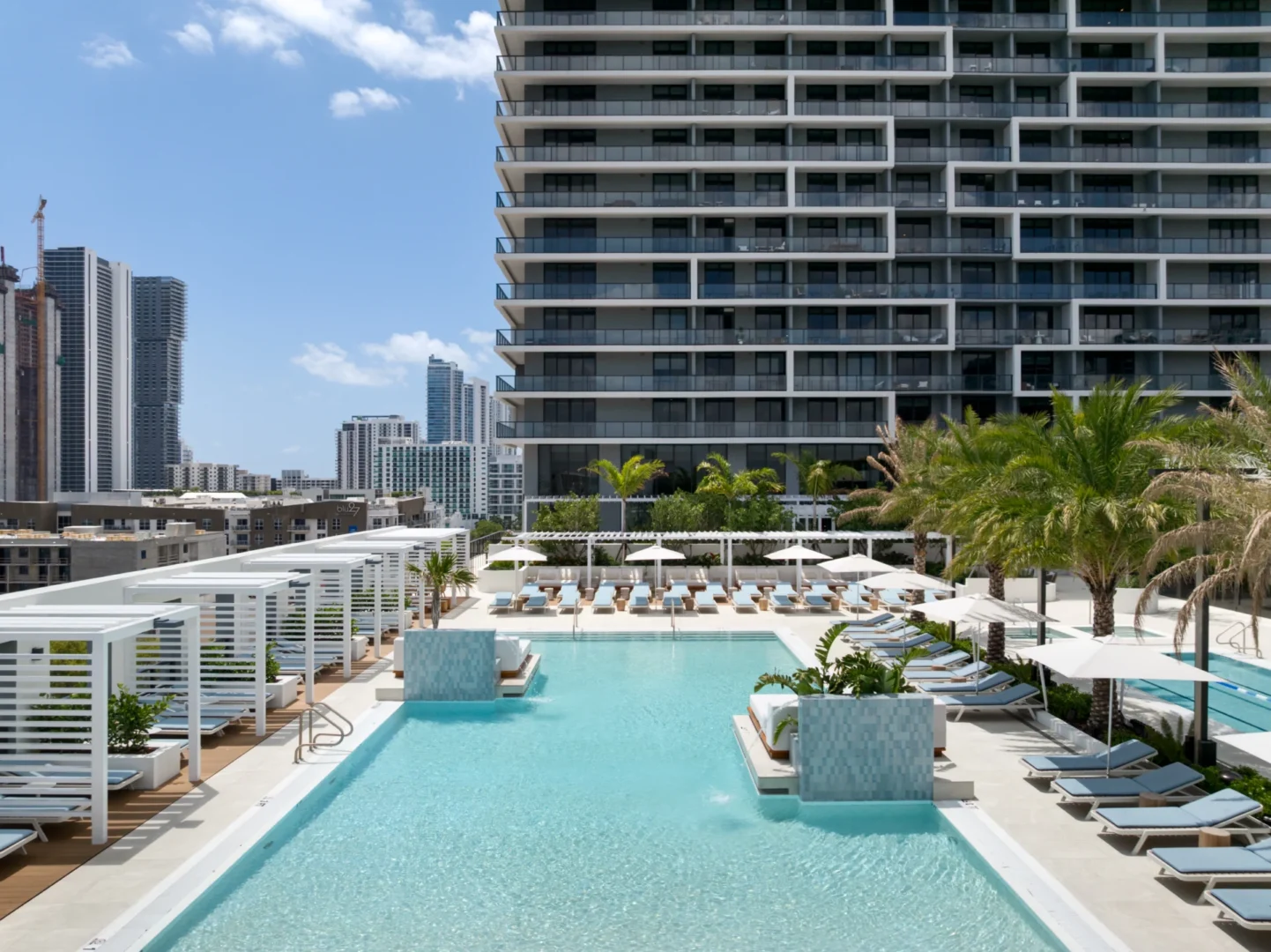 A multi-story building in the Forma project stands behind a swimming pool terrace with lounge chairs, umbrellas, and white cabanas