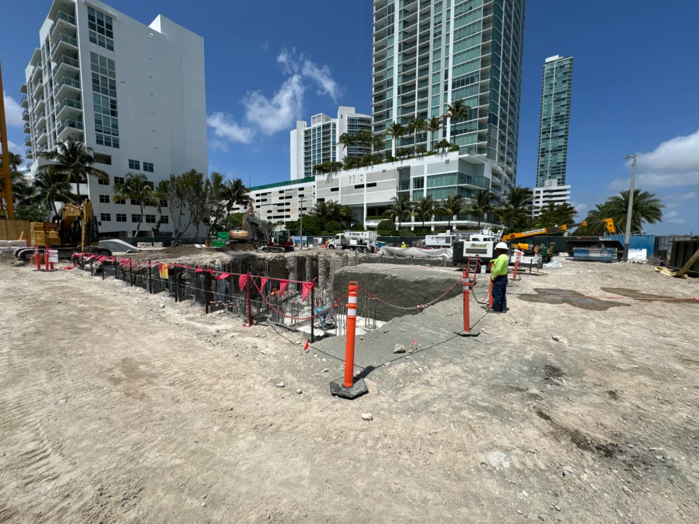 An excavated construction site at Villa Miami contains heavy machinery, a worker, and tall buildings nearby