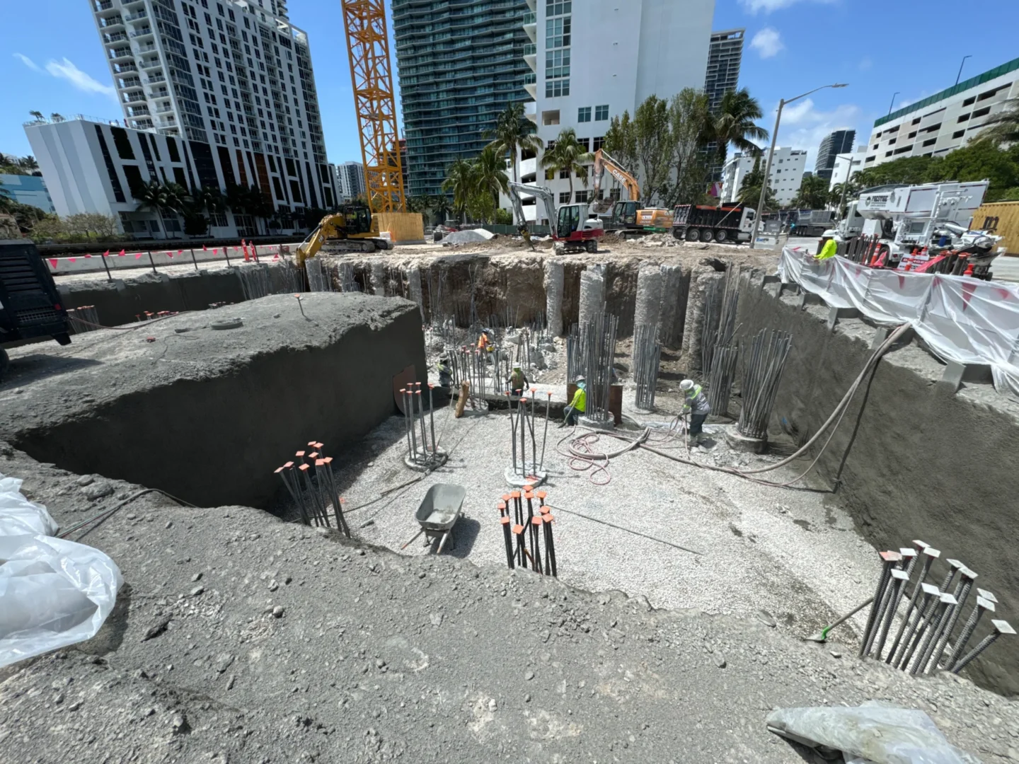 An excavation pit for Villa Miami contains heavy machinery, structural rebar, and workers near high-rise buildings