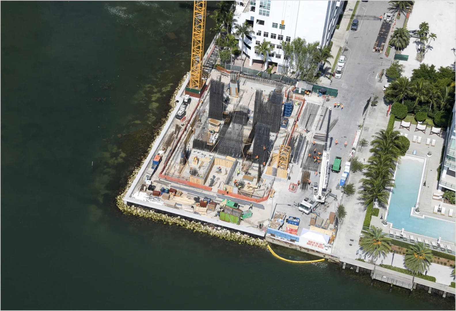 Rebar structures and a yellow crane at the Villa Miami construction site sit along the edge of dark green water