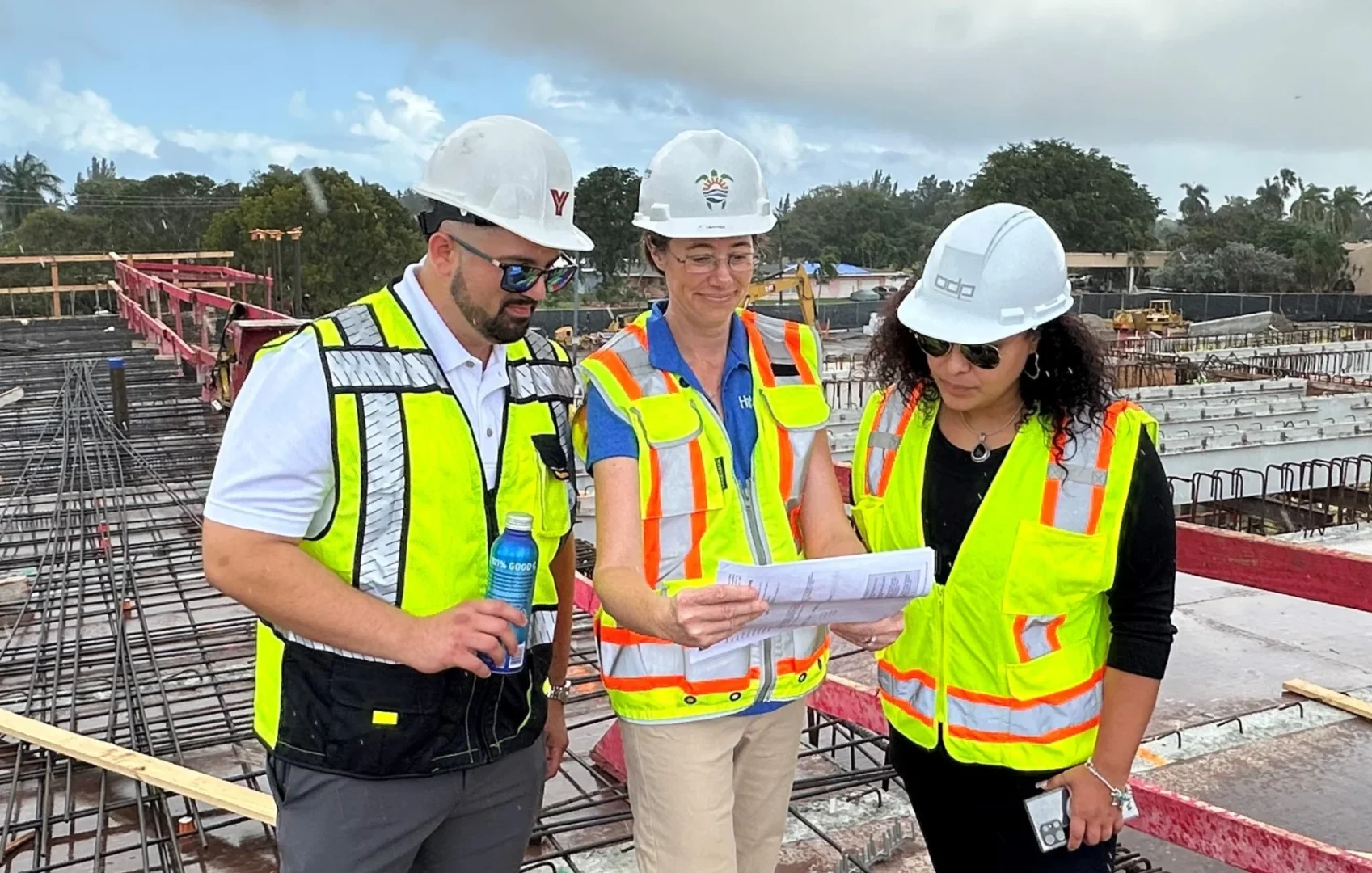 Three individuals wearing high-visibility vests and hard hats review papers at the Hollywood Police Headquarters construction site
