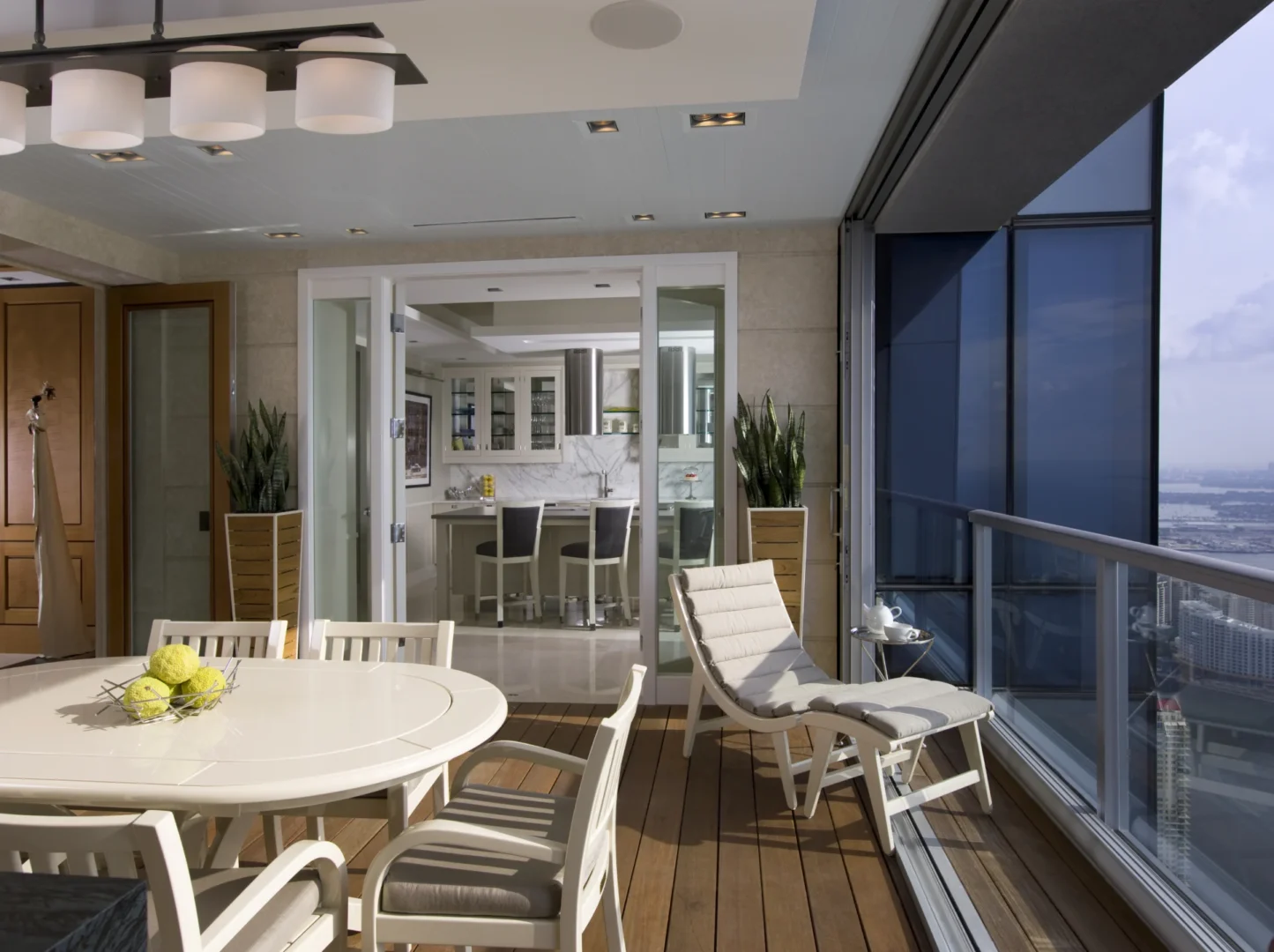 White dining furniture and a lounge chair sit on the wooden terrace of Penthouse 4 next to an indoor kitchen