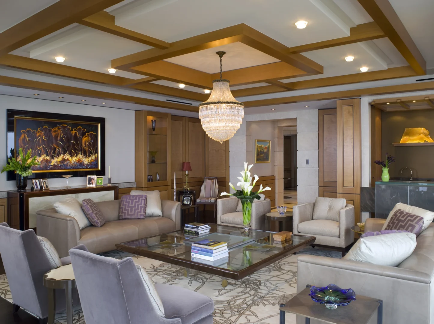 A crystal chandelier hangs from a coffered ceiling in the Penthouse 4 living room with beige seating around a glass coffee table