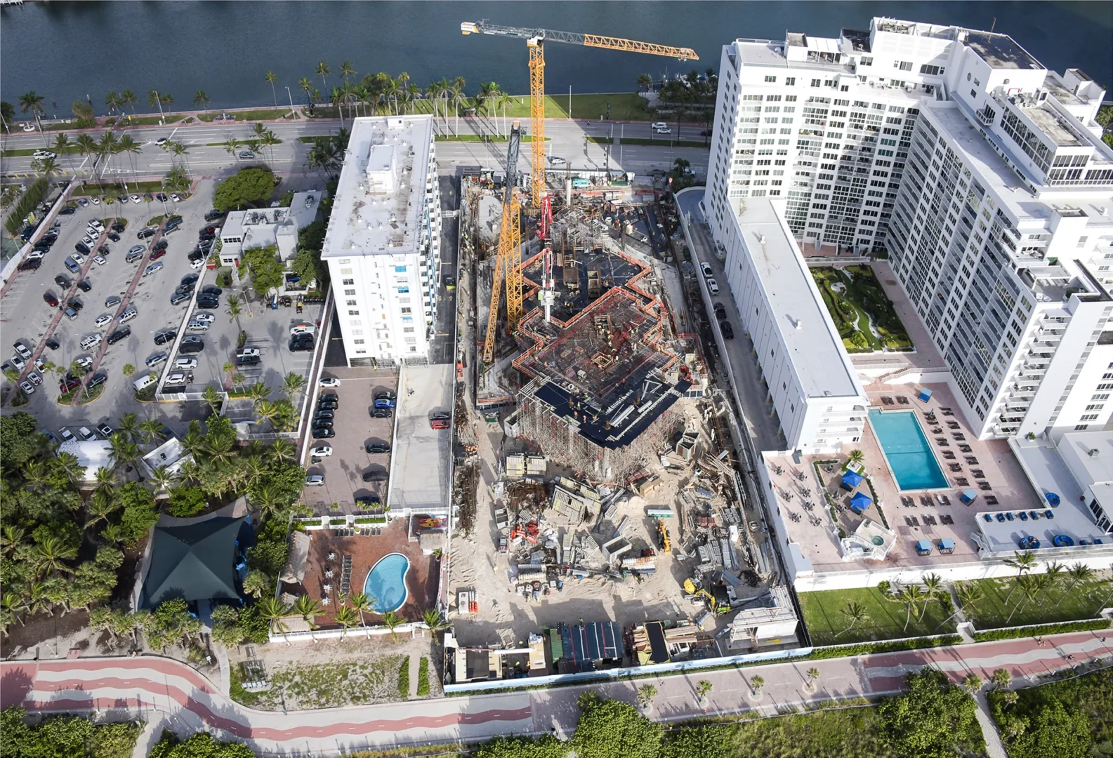 Construction cranes and frameworks for The Perigon are situated between high-rise buildings, parking lots, and a body of water
