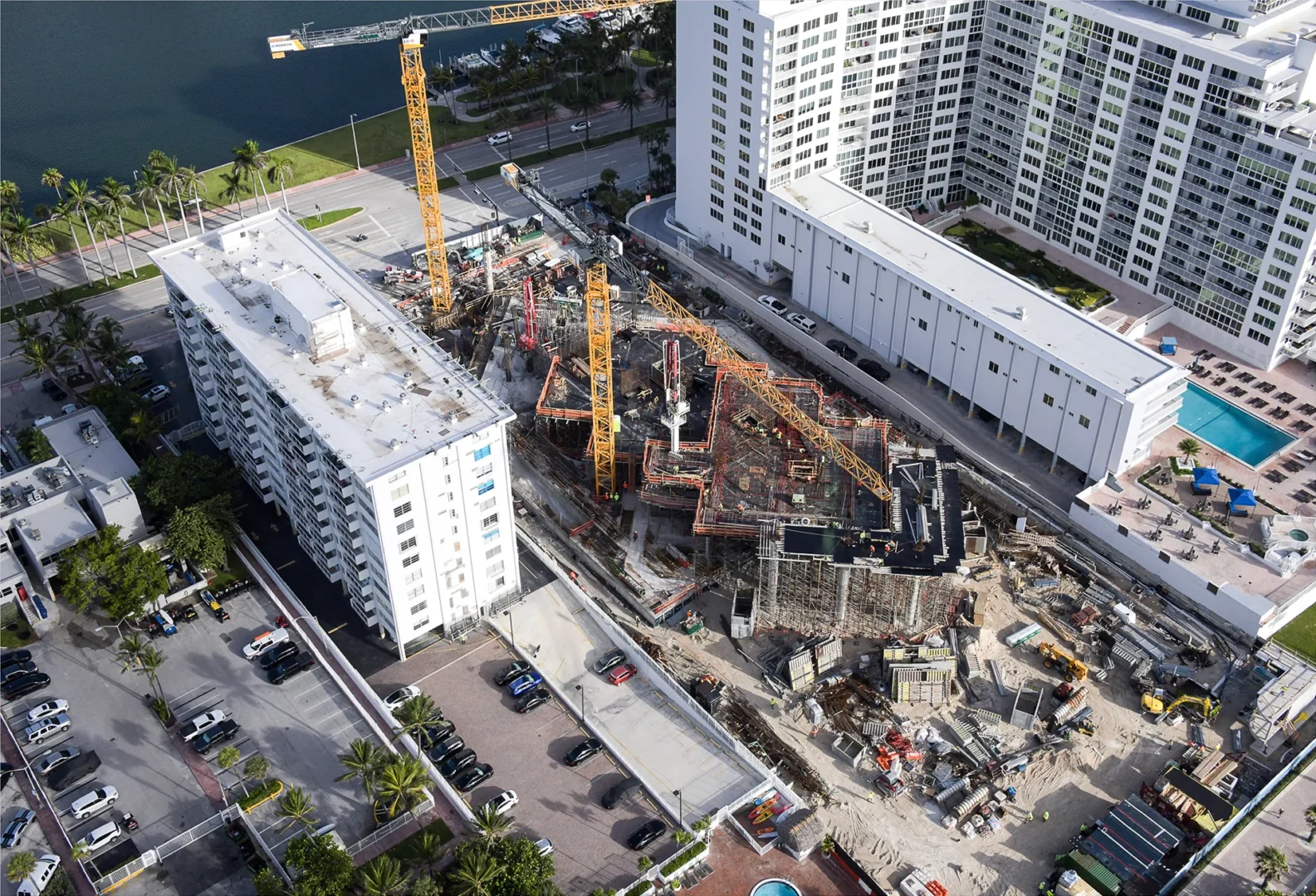 Yellow cranes tower over the construction site for The Perigon, positioned between large white buildings and a roadway near water