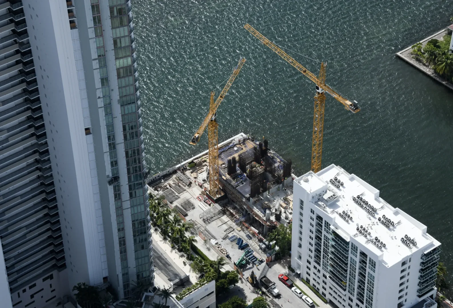 Two yellow cranes at the Villa Miami construction site stand between tall buildings and a body of water