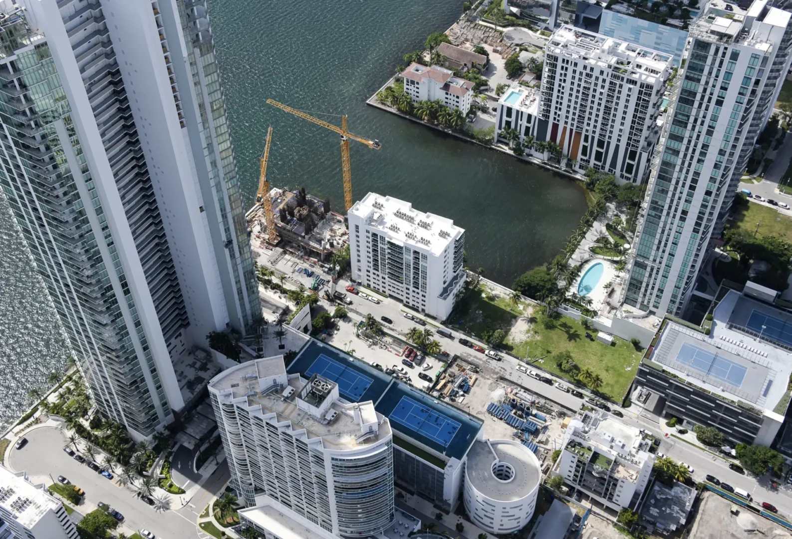 Tall white skyscrapers and a construction site with yellow cranes border a waterway near Villa Miami