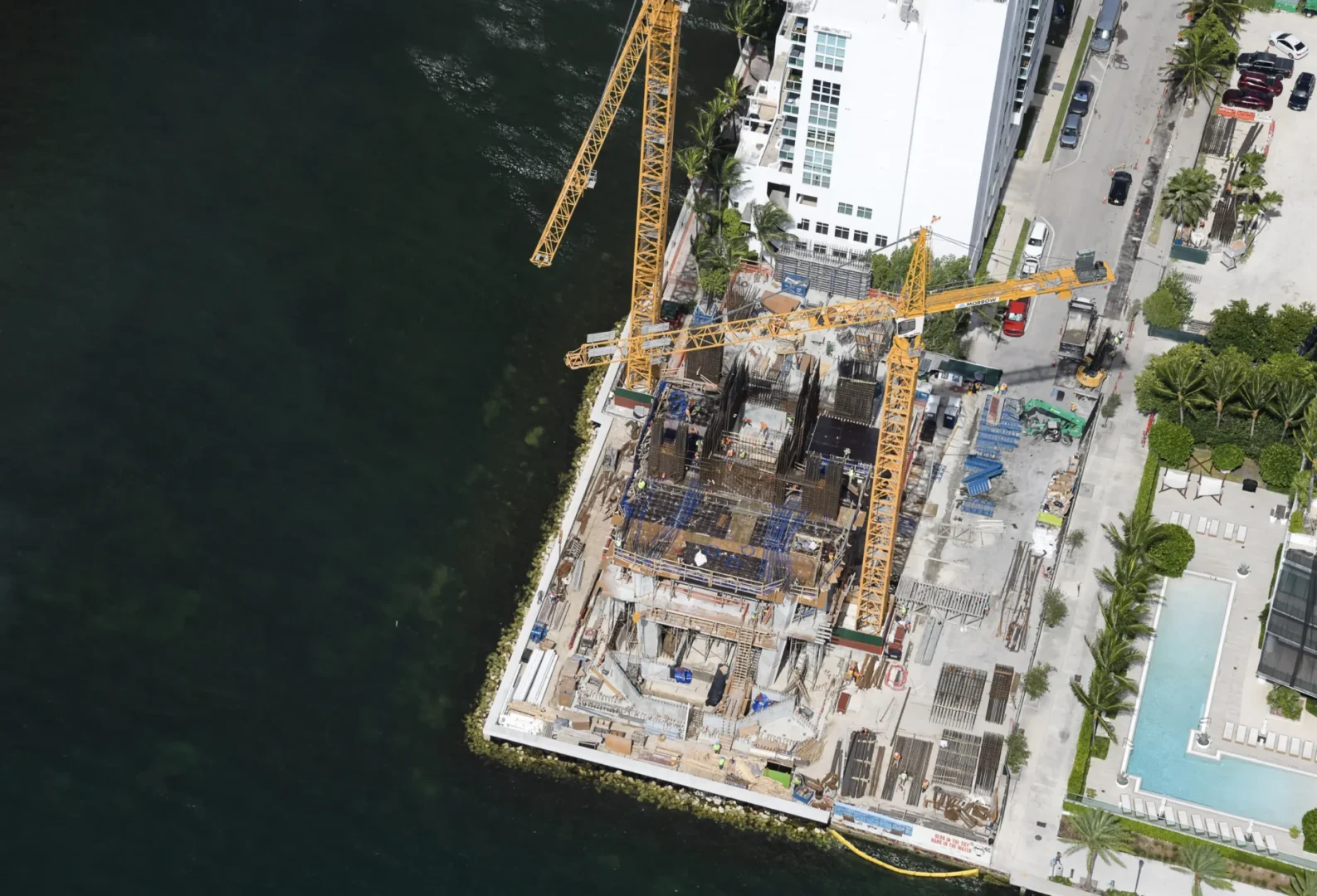 Two yellow cranes stand over a construction site for Villa Miami next to dark water, white buildings, and a swimming pool